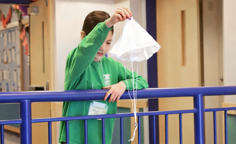 Boy in a green shirt is standing behind a metal rail, preparing to drop a white parachute over the railing.